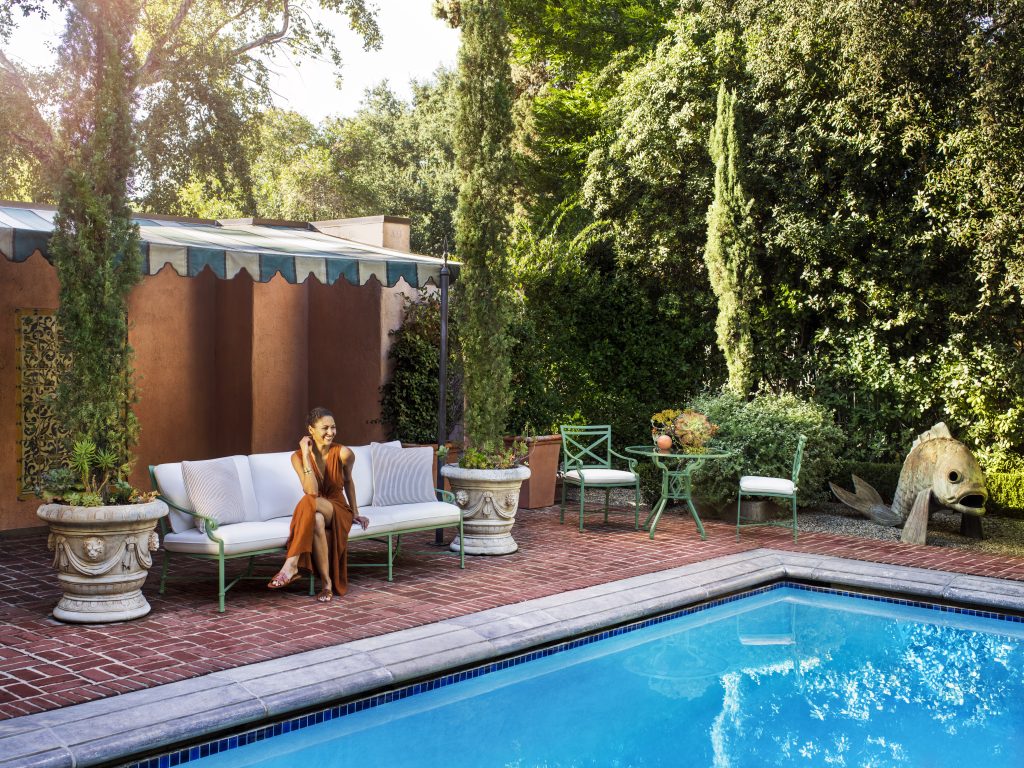 A woman seated on an outdoor sofa next to a pool with beautiful trees and plants