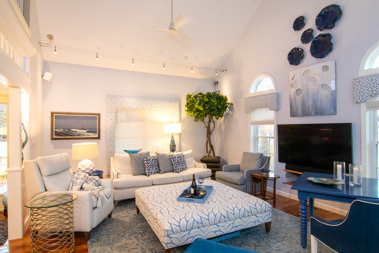 A living room with vaulted ceiling, white furniture and different shades blue accents. There is a large TV, a potted tree and dark blue ceramic plates as decor on the walls. 