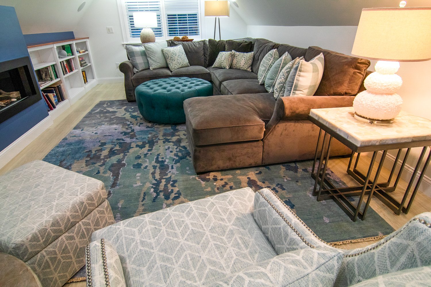 A comfy den with a deep soft couch, round teal leather ottoman, and blue rug